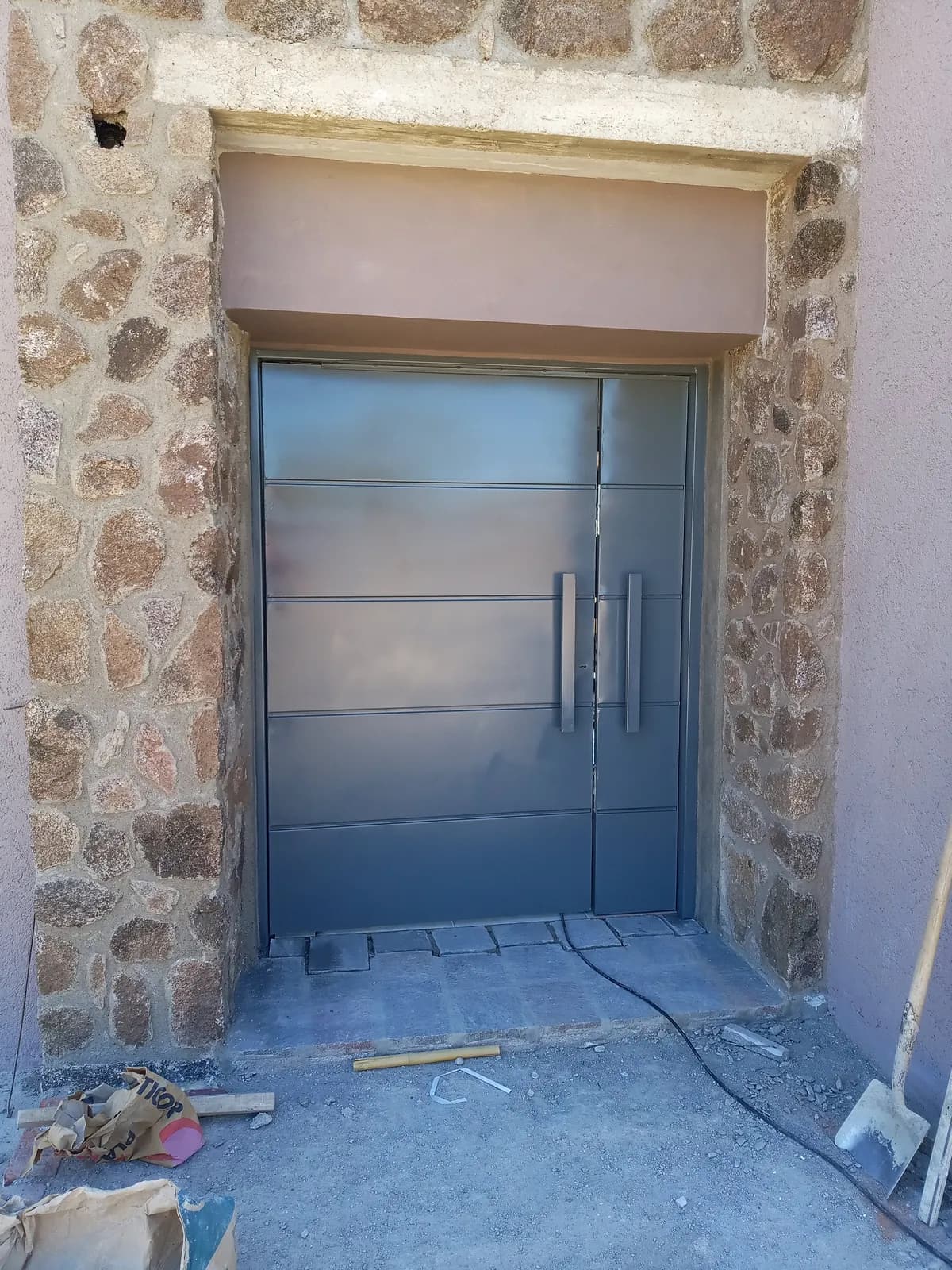 Puerta de chapa gris con líneas horizontales y barrales, en un ingreso de piedra